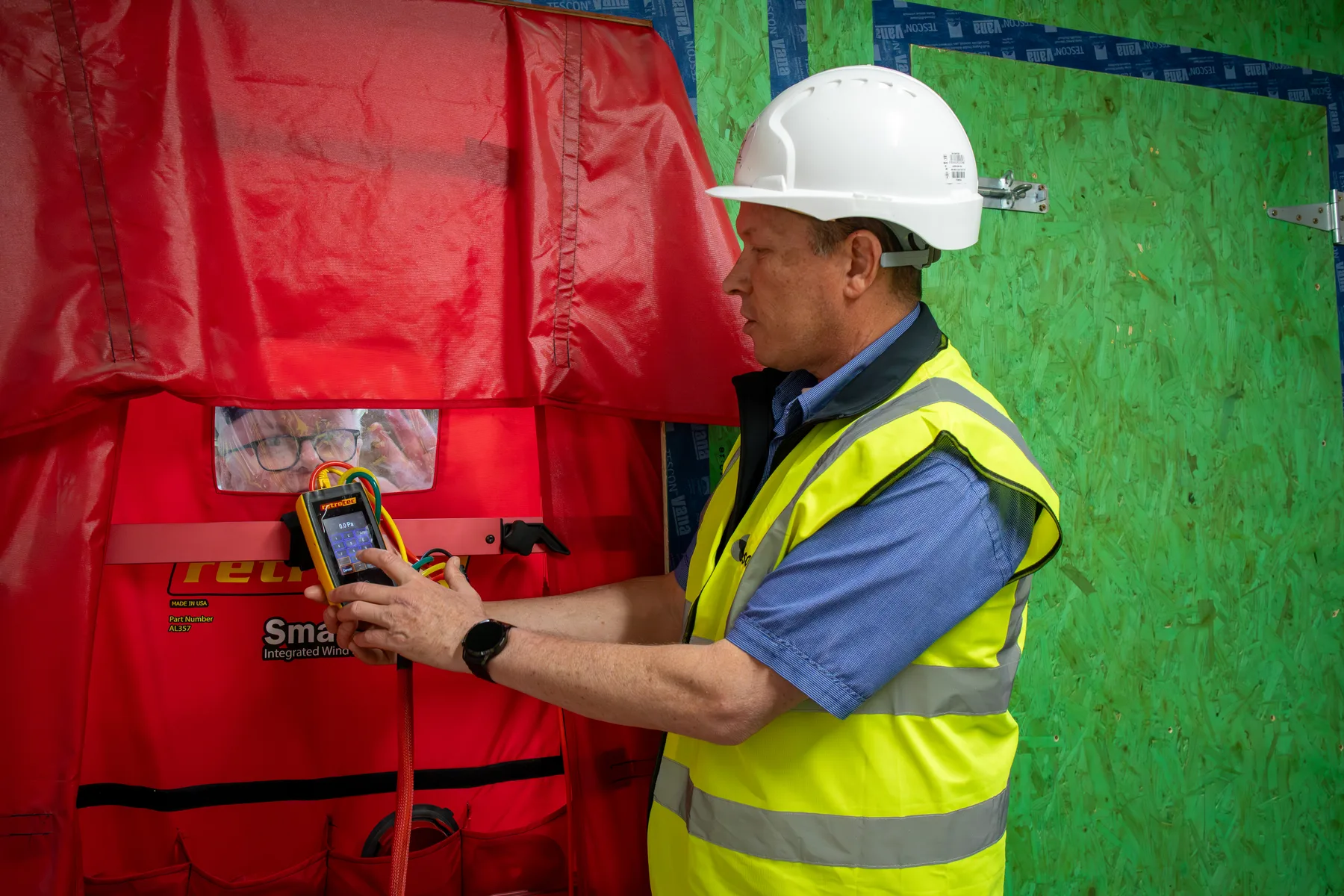 A blower-door airtightness test in progress: a tester in hi-vis and hard hat reading the manometer at the red door shroud, with the taped airtight layer behind